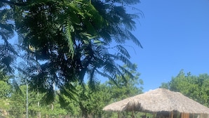 Una estructura con techo de paja y asientos al aire libre, rodeada por un área de grava y follaje.