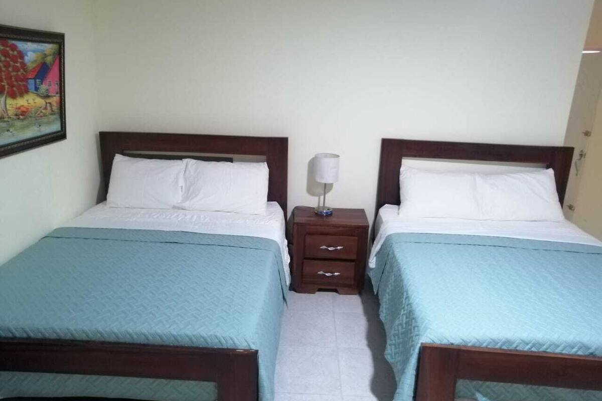 Una habitación con dos camas individuales, cada una con cabecero de madera y colcha azul claro. Entre las camas, una mesita de noche de madera con una lámpara.