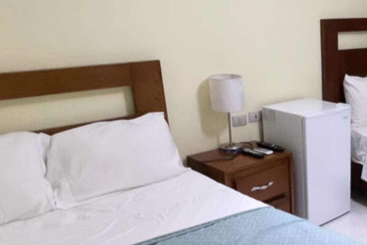 Habitación de hotel con cama, mesita de noche, lámpara y un pequeño electrodoméstico blanco.