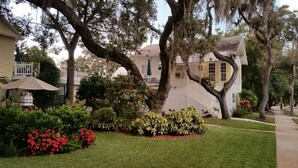 Une grande maison blanche dotée d’un porche à l’avant, entourée d’une végétation luxuriante et de plantes en fleurs.