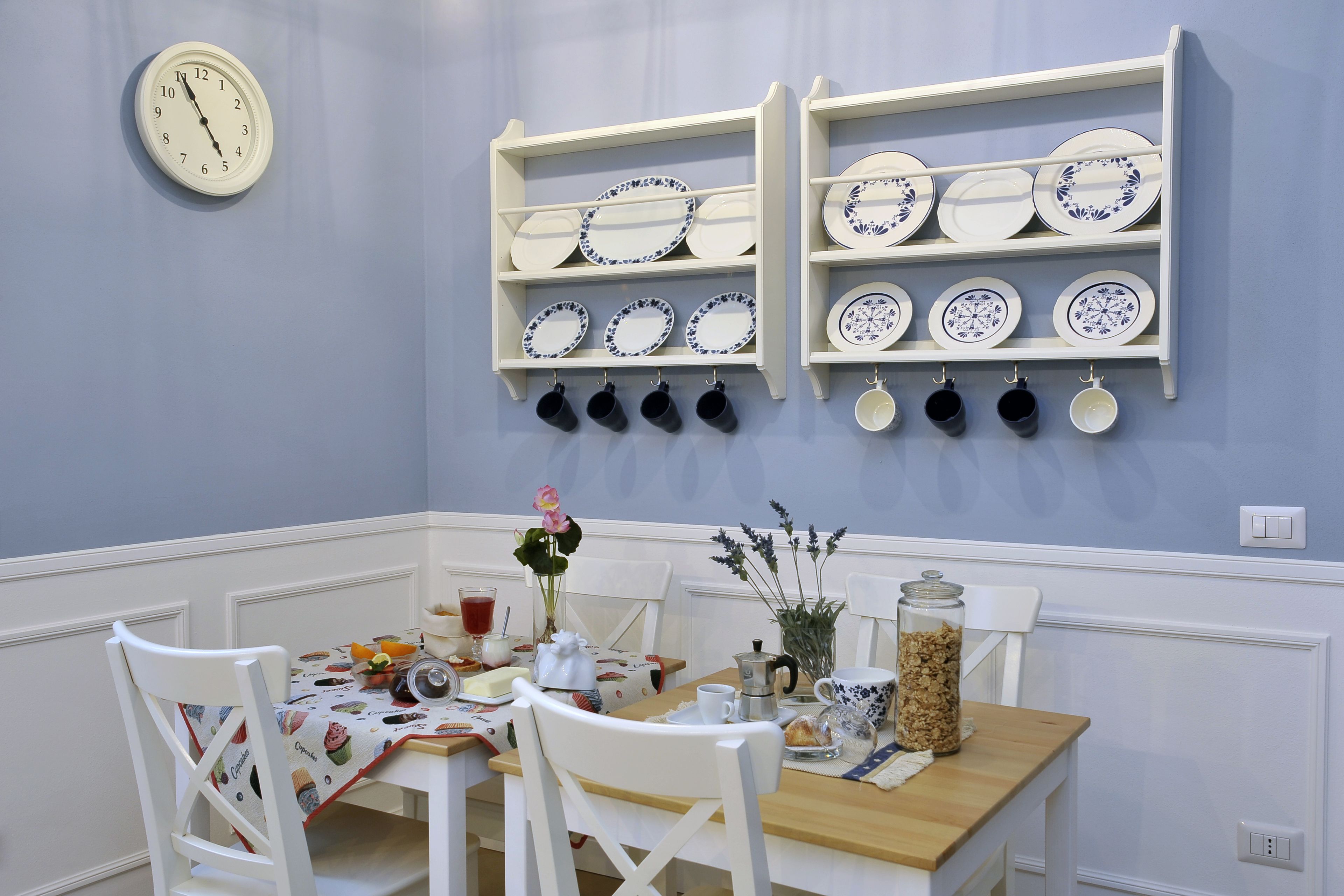 Un comedor con una mesa dispuesta para dos, un reloj en la pared y estantes con platos y tazas.