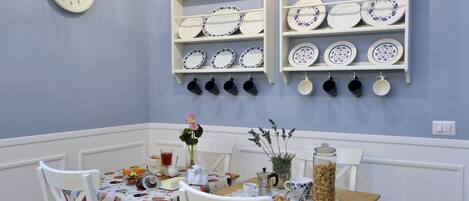 Un comedor con una mesa dispuesta para dos, un reloj en la pared y estantes con platos y tazas.