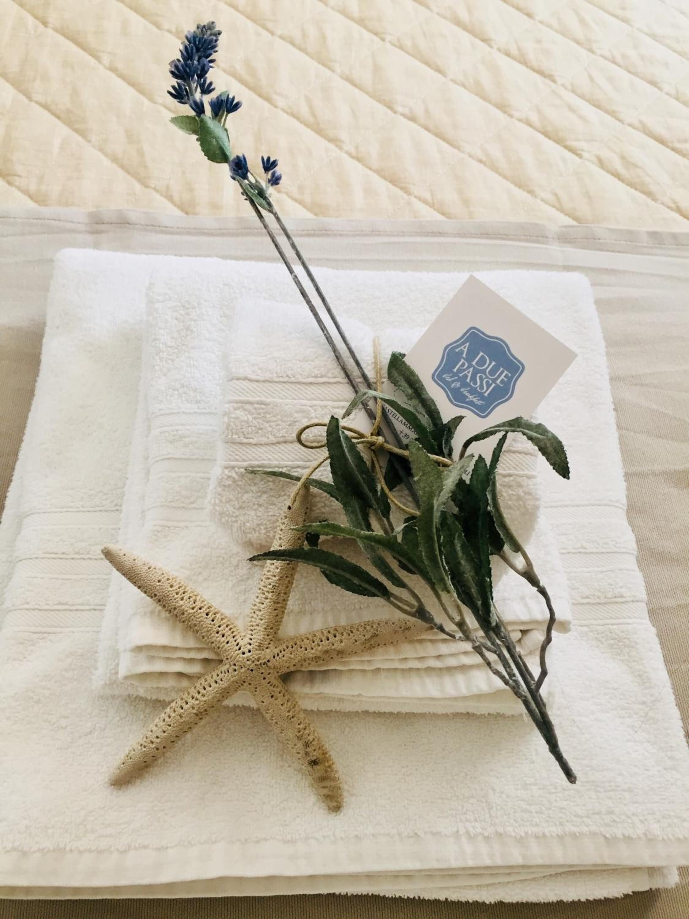 A neatly folded white towel with a tag labeled "A DUE PASSI" and a dried lavender branch with a starfish.
