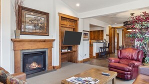 Een woonkamer met een open haard, een televisie, een bank, een salontafel en een eethoek met houten kasten.