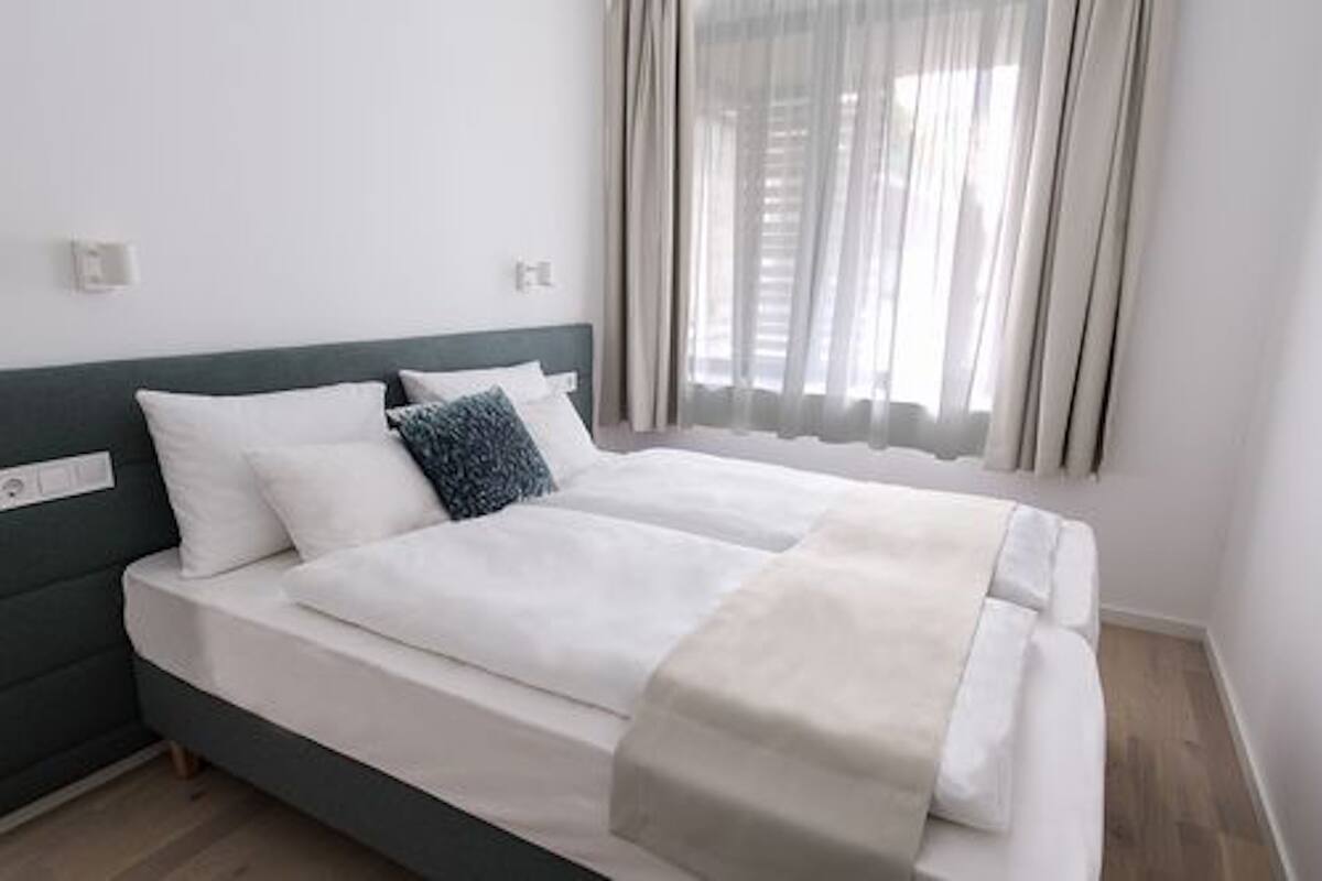 Ein Doppelbett mit weißer und grauer Bettwäsche, ein grüner Kopfteil und ein Fenster mit durchscheinenden Vorhängen.