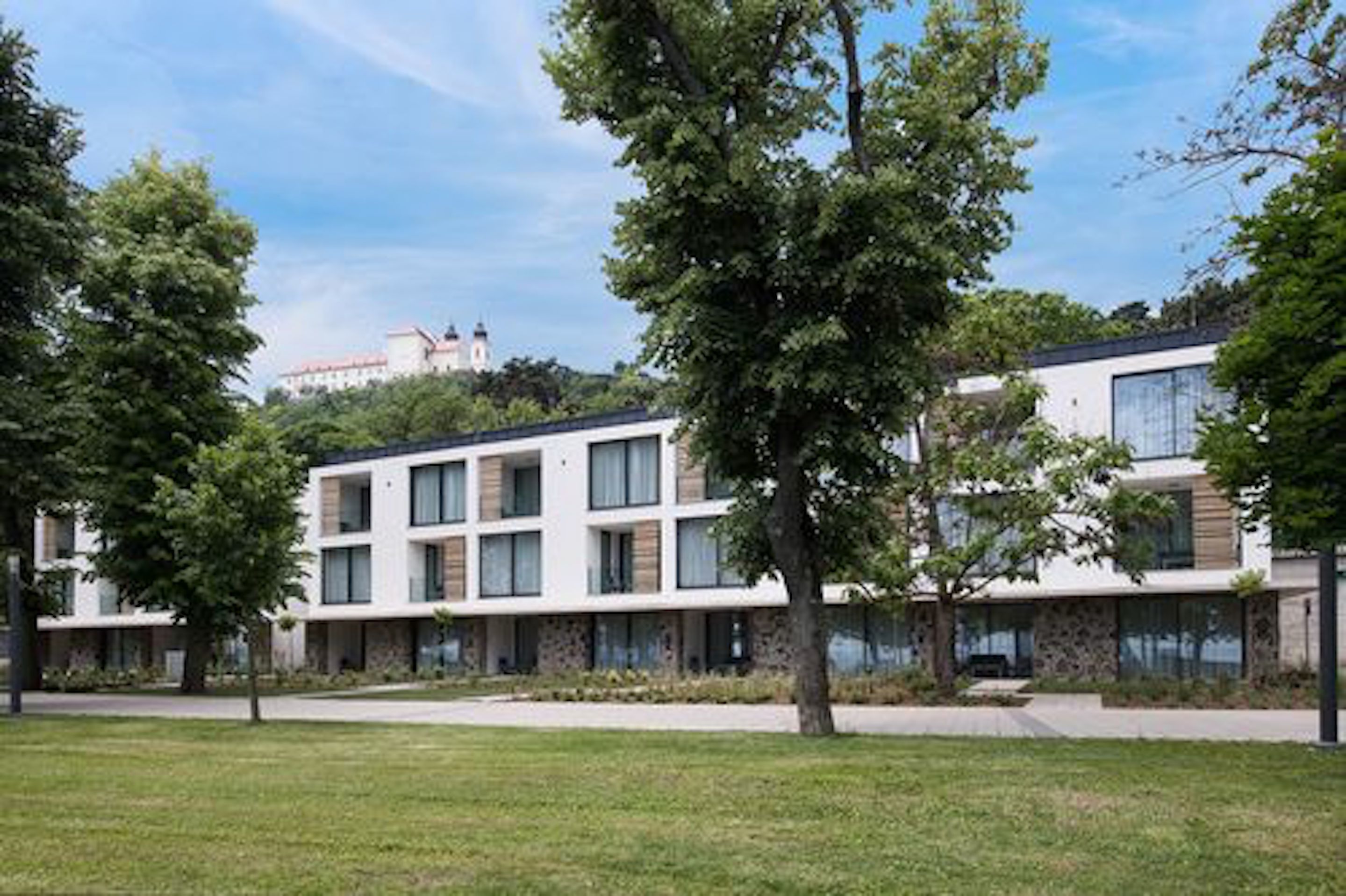 Um edifício moderno com telhado plano, janelas amplas e fachada de pedra. Árvores estão em frente ao edifício.