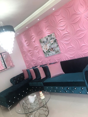 A modern living room with a pink textured wall, a floral painting, a dark blue sofa with teal cushions, and a glass coffee table with a white object on it.