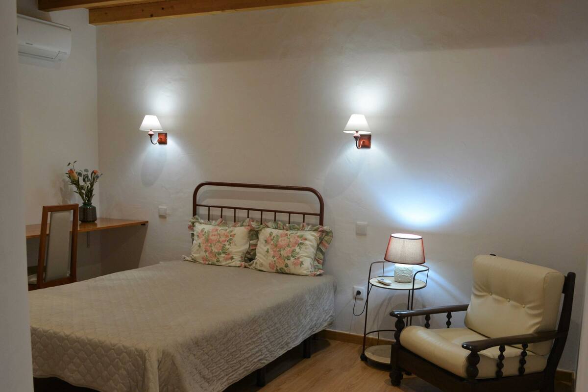 A bedroom with a bed, a chair, a nightstand, and two wall-mounted lamps.