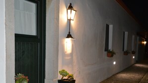 Une ruelle étroite pavée, bordée d’un bâtiment blanc avec une porte verte et une lampe fixée au mur, illuminée.