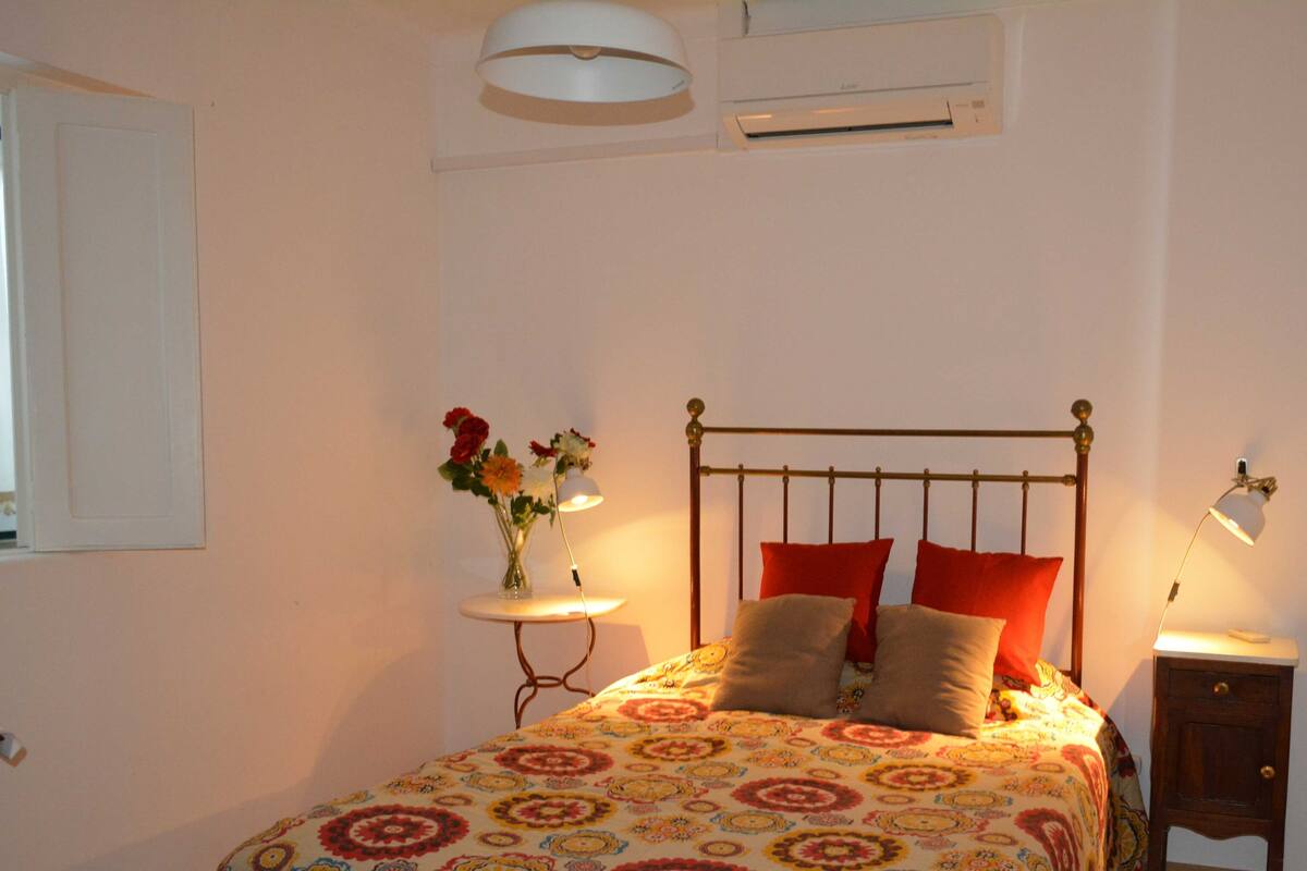 A bedroom with a bed, a bedside table, a lamp, a wall-mounted air conditioner, and a window with curtains.
