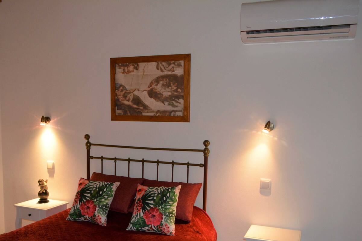 A bedroom with a bed, two bedside tables, a wall-mounted air conditioner, and a framed picture above the bed.