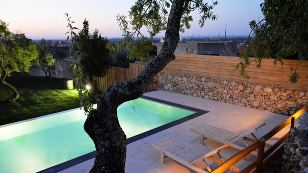 Une piscine entourée de chaises longues et dotée d’une terrasse en bois, le tout bordé d’un mur de pierre et d’arbres.