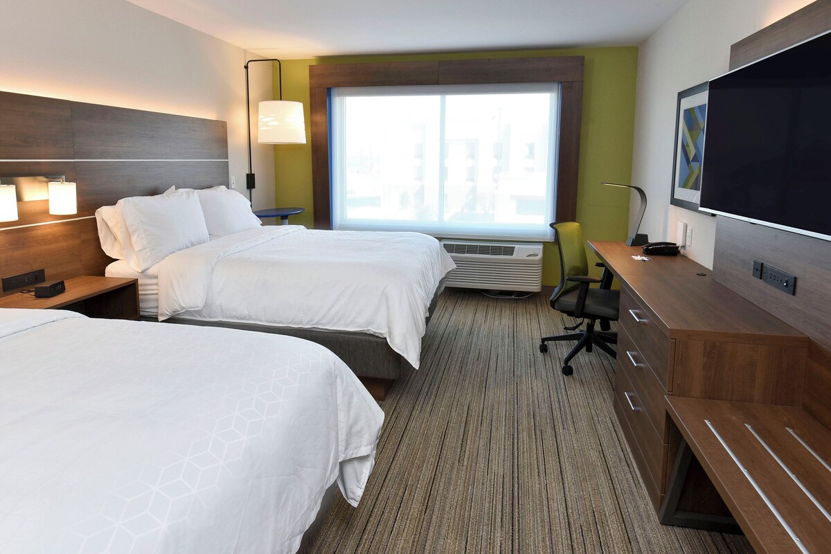 A hotel room with two beds, a desk, a flat-screen TV, a lamp, and a window with a view of buildings.