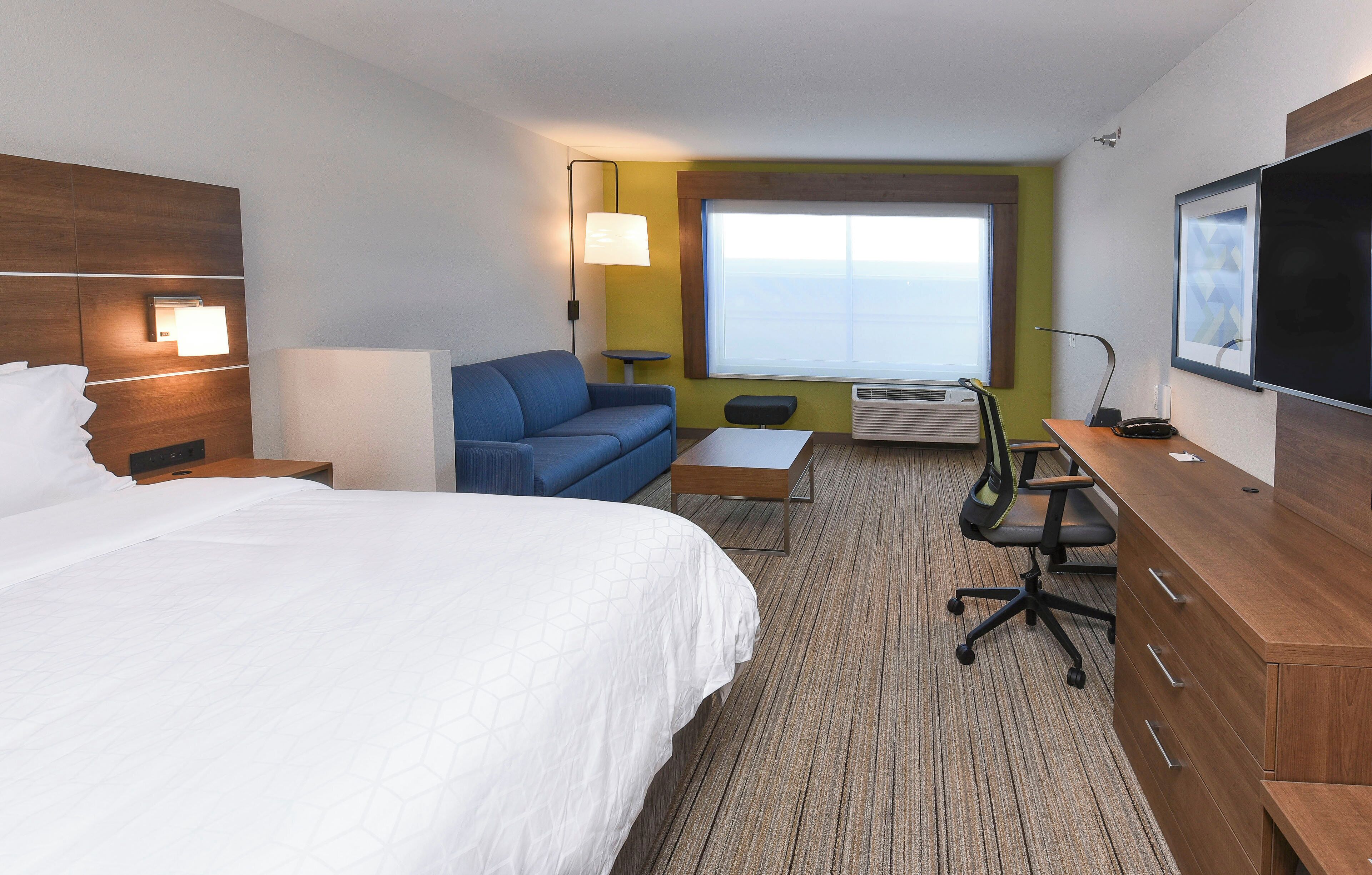 Quarto de hotel com uma cama grande, um sofá azul, uma mesa pequena, uma escrivaninha com cadeira, televisão e janela com vista.