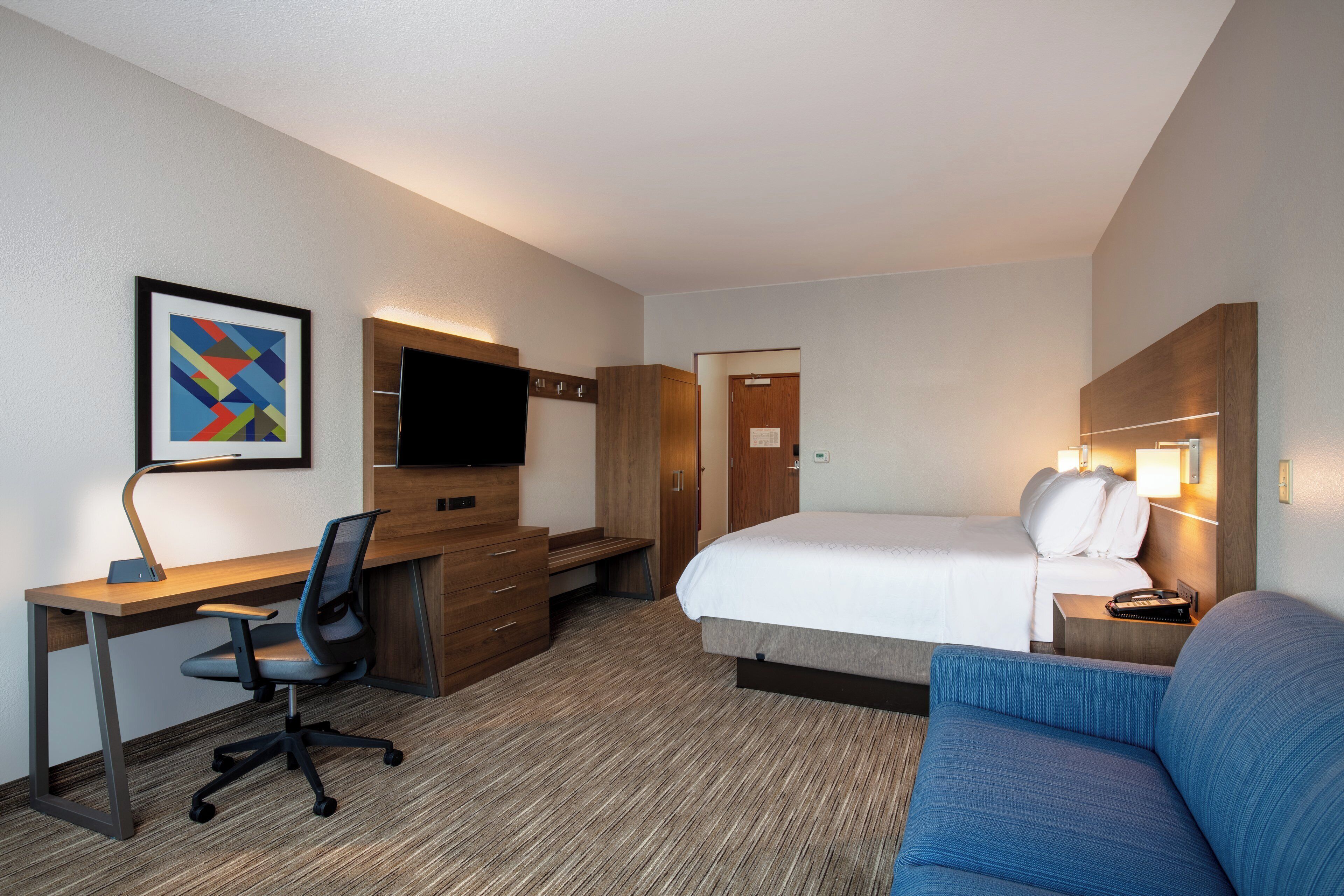 Quarto de hotel com cama, escrivaninha com cadeira, televisão, sofá azul e guarda-roupa de madeira.