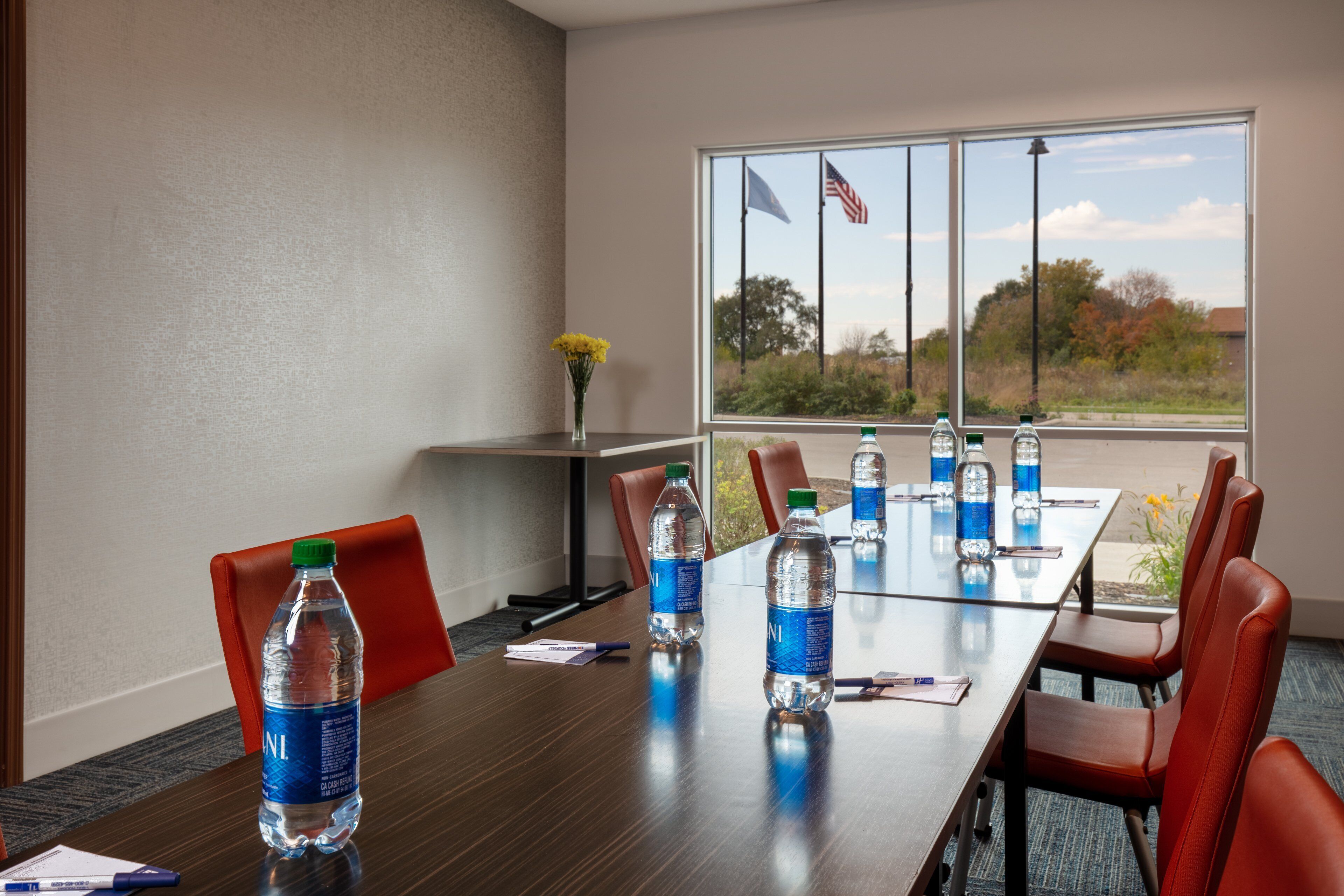 A conference room with a long table, red chairs, water bottles, and a vase with yellow flowers.