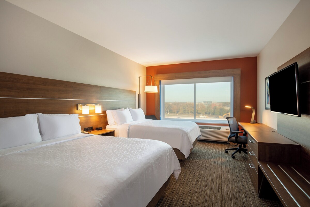 A hotel room with two beds, a desk, a chair, a TV, and a large window.