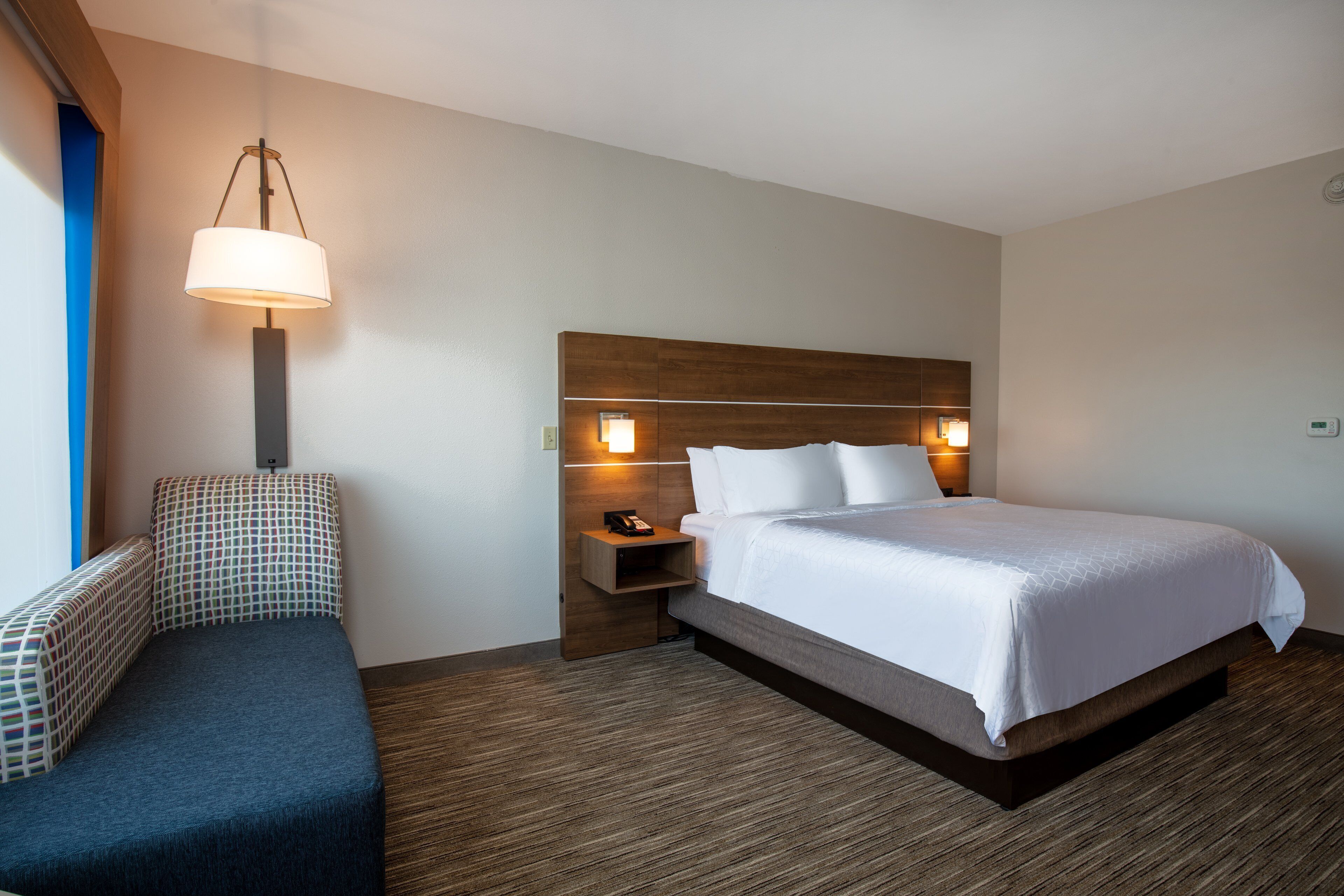 A hotel room with a bed, bedside table, lamp, and a bench.