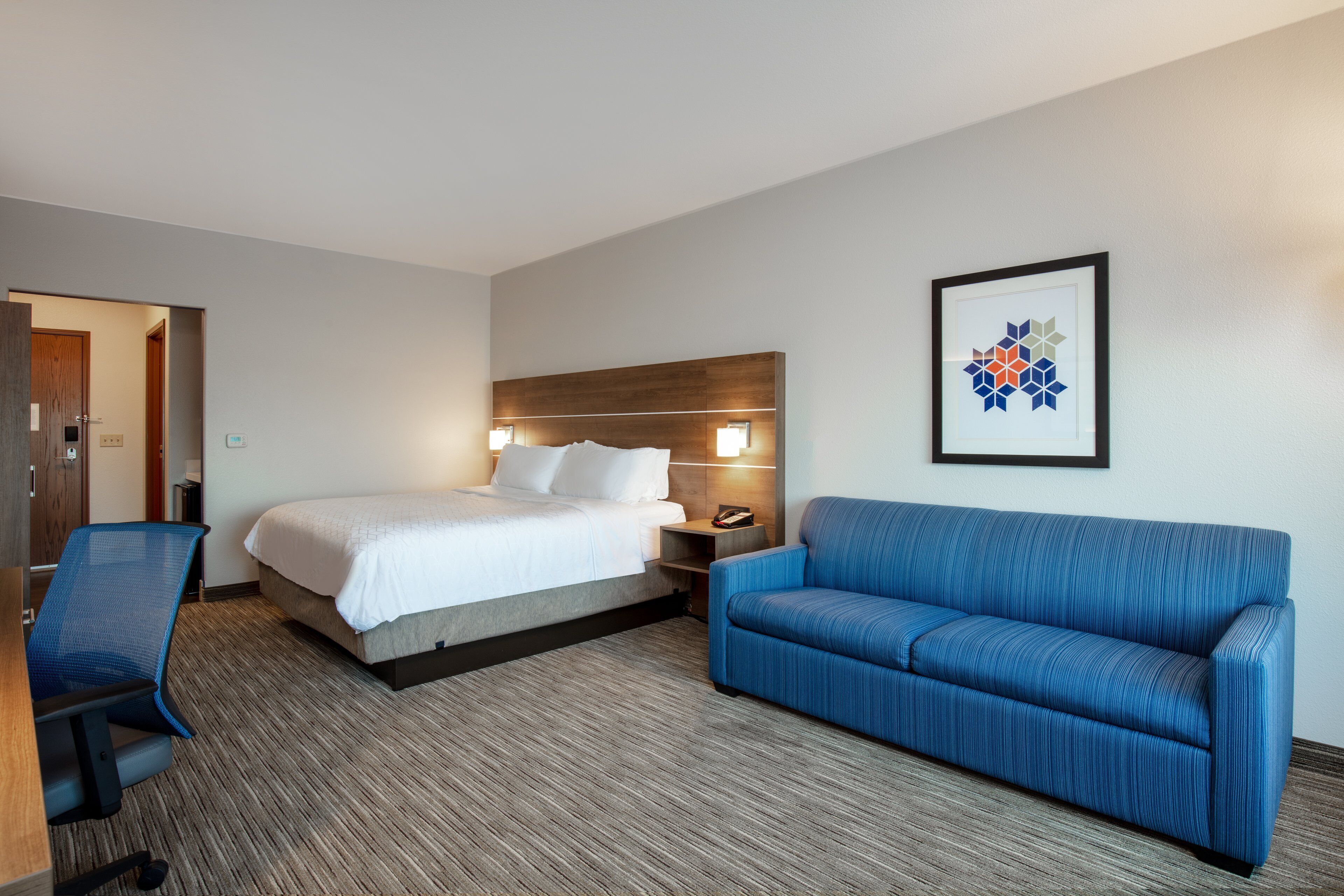 A hotel room with a bed, a blue sofa, a desk with a chair, and a framed picture on the wall.