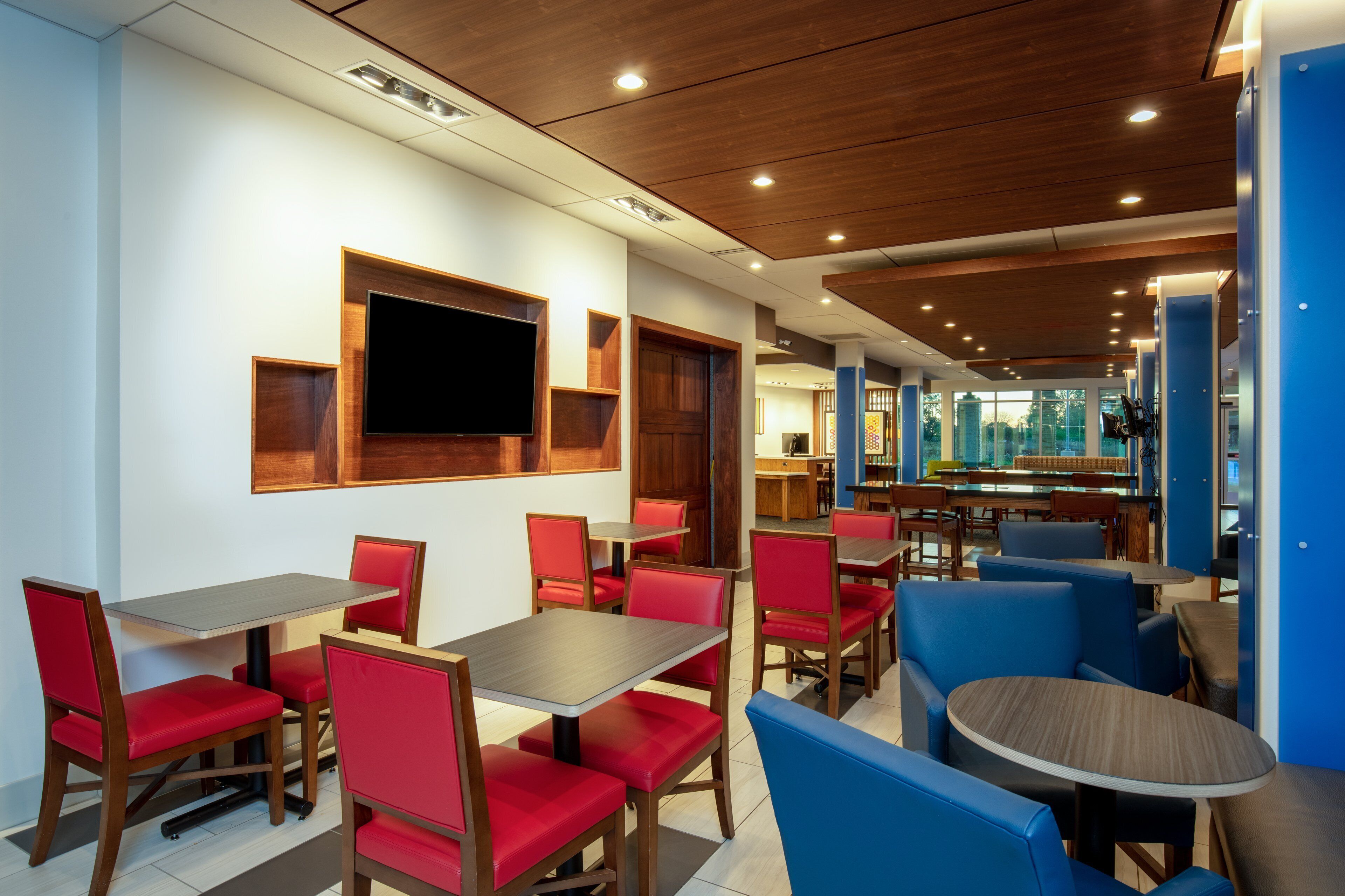 Un restaurant moderne avec des panneaux de plafond en bois, des chaises recouvertes d’un tissu rouge et bleu, et un espace de restauration central doté d’un grand écran de télévision.
