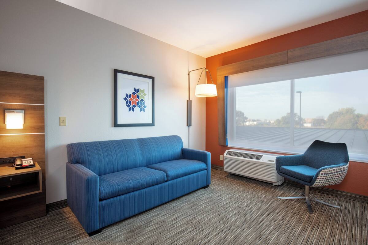 A hotel room with a blue sofa, a chair, a framed artwork, a lamp, and a large window.