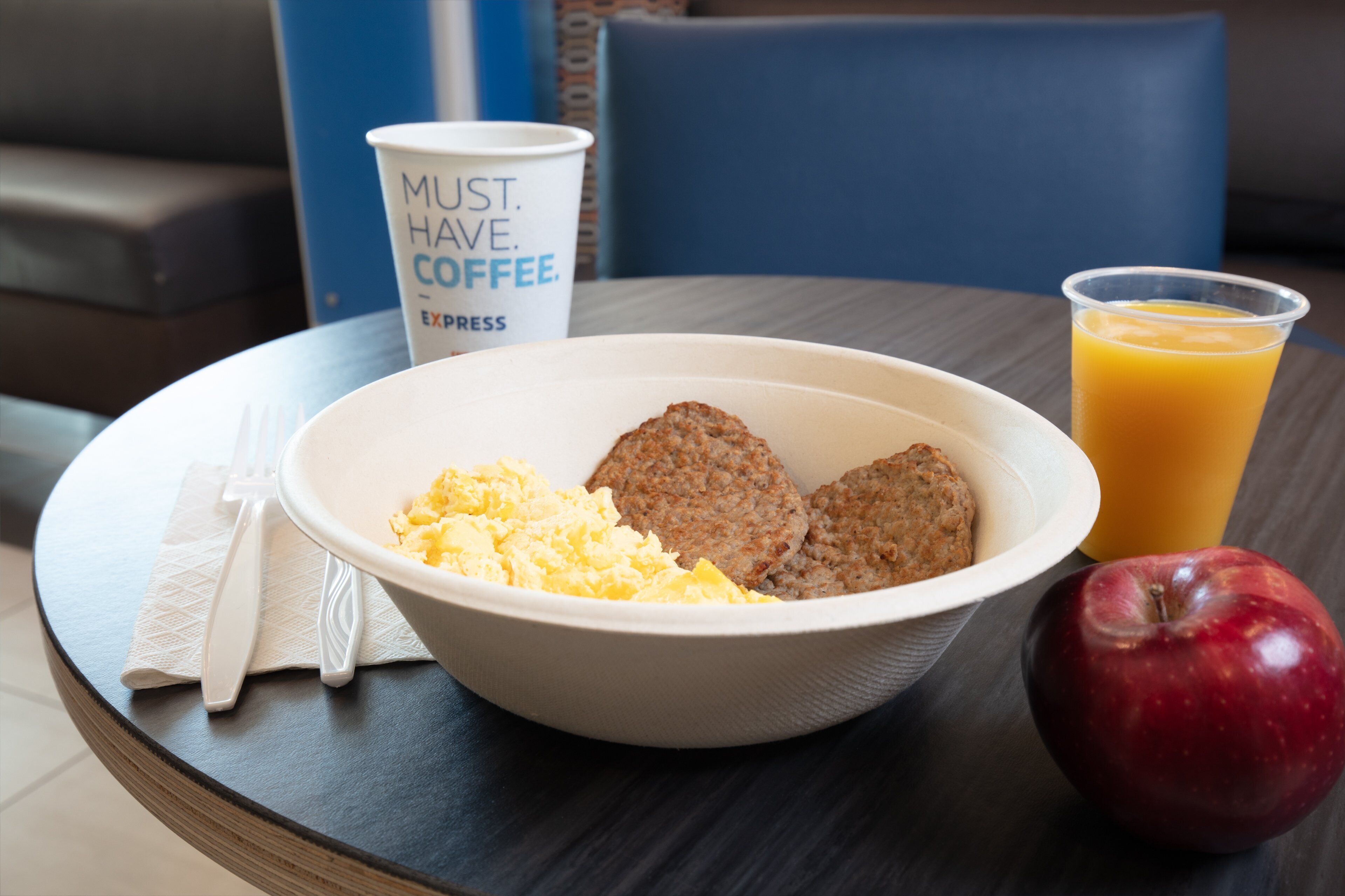 A breakfast tray with scrambled eggs, sausage patties, a red apple, a cup of coffee, and a glass of orange juice on a round table.