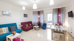 Una sala de estar con un sofá azul, una pequeña mesa blanca con un florero, un sillón rojo, una mesa de comedor de madera con sillas, y un televisor en la pared.