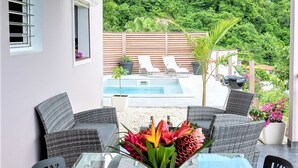Une table à manger pour quatre personnes, avec un plateau en verre, entourée de chaises en osier, et donnant sur une piscine et une végétation luxuriante en arrière-plan.