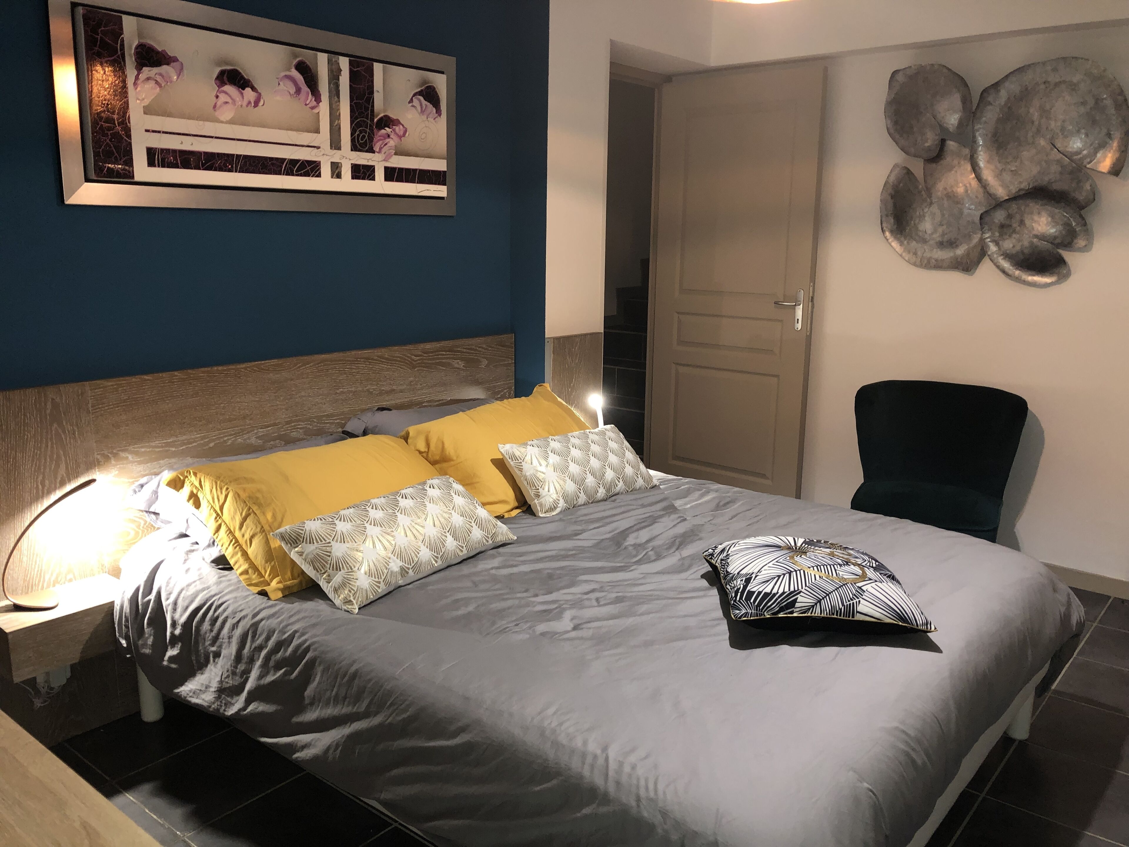 Un lit bien fait, recouvert d’une couvre-lit grise et jaune, agrémenté d’oreillers blancs à motifs et d’un coussin noir et blanc. On aperçoit une tête de lit en bois, une table de chevet avec une lampe, et une œuvre d’art murale au-dessus du lit.