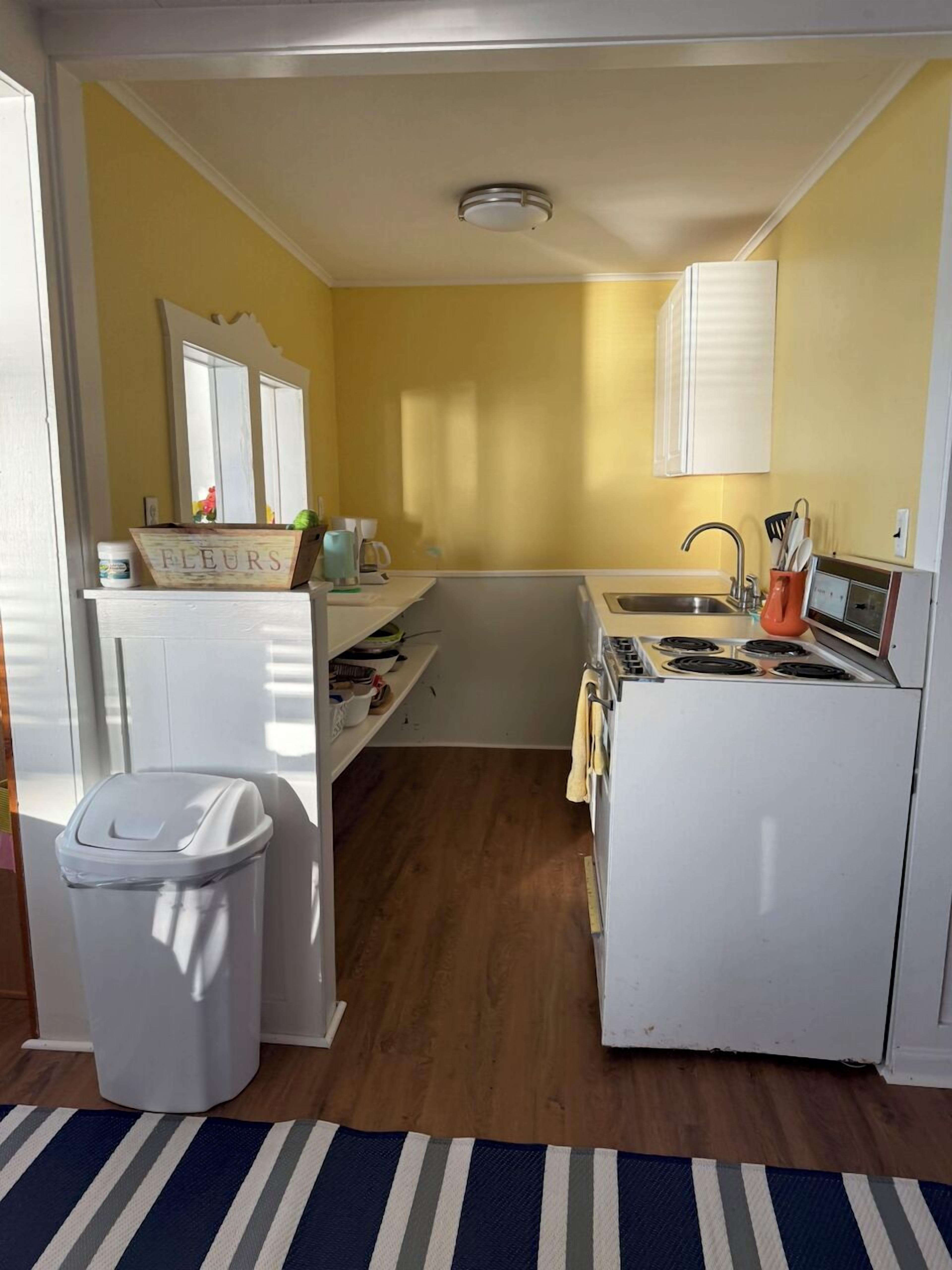 Une petite cuisine avec un réfrigérateur, une cuisinière et un évier blancs. On y trouve une poubelle et une boîte en bois étiquetée « FLEURS » sur le plan de travail.