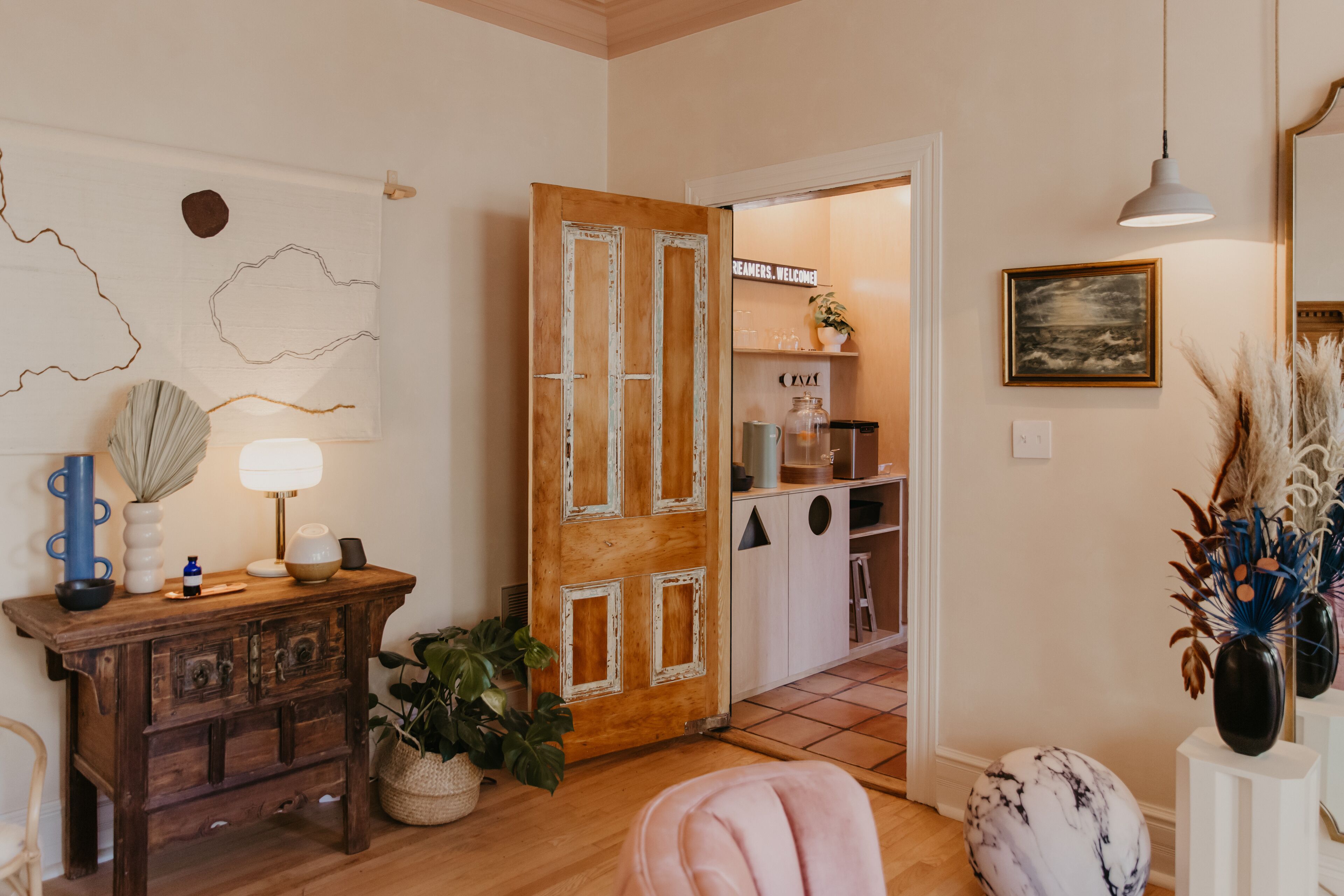 Quarto acolhedor com porta de madeira, parede branca com arte abstrata, pequena mesa com abajur e planta em vaso.