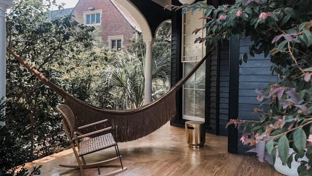 A hammock chair in a room with a wooden floor, a small trash bin, and a view of a house through an arched window. - Dreamers BNB (Wilmington)