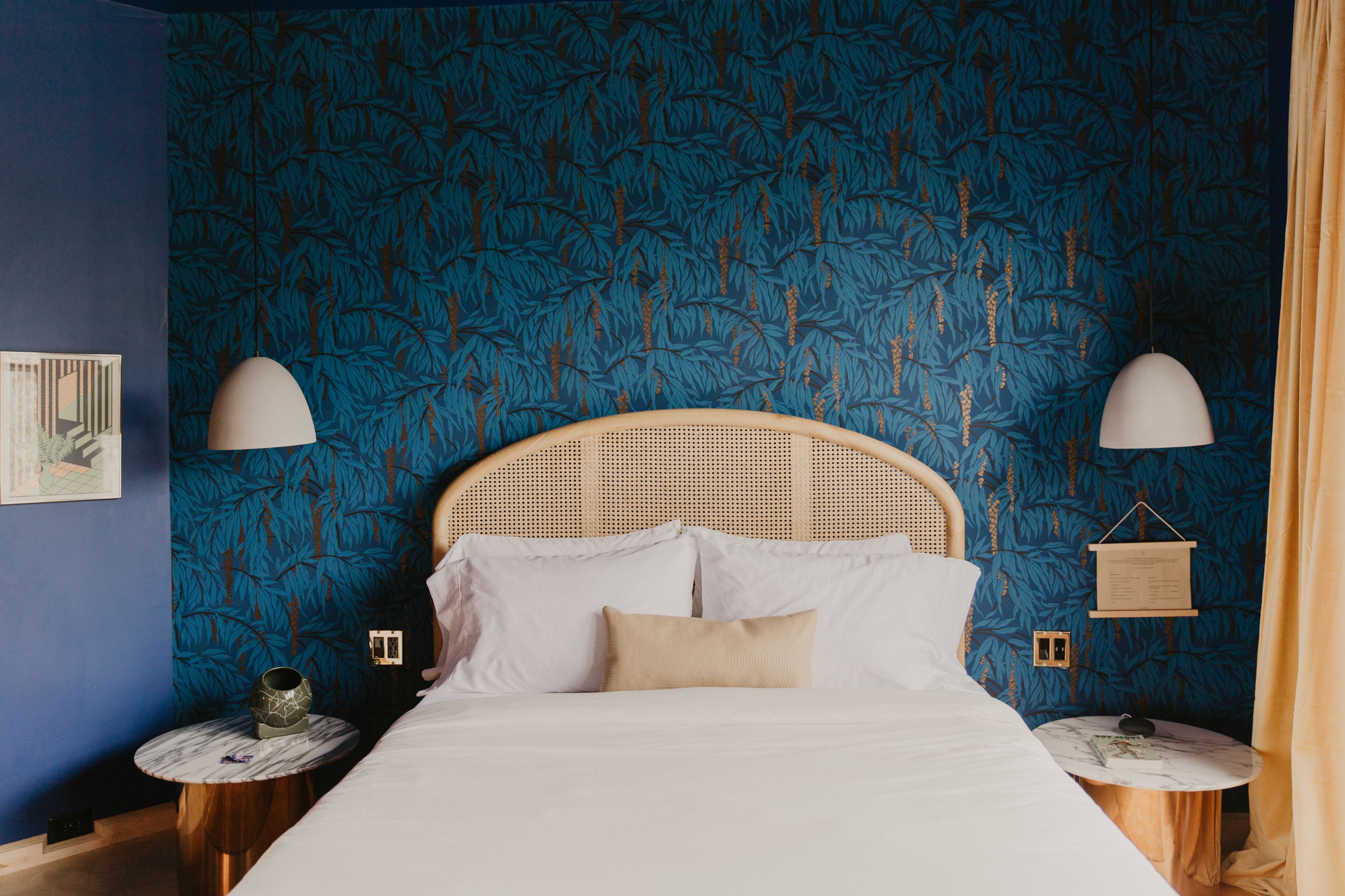 A bedroom with a rattan headboard, white bedding, and a blue leaf-patterned wallpaper. There are two bedside tables with lamps and a small wooden shelf.