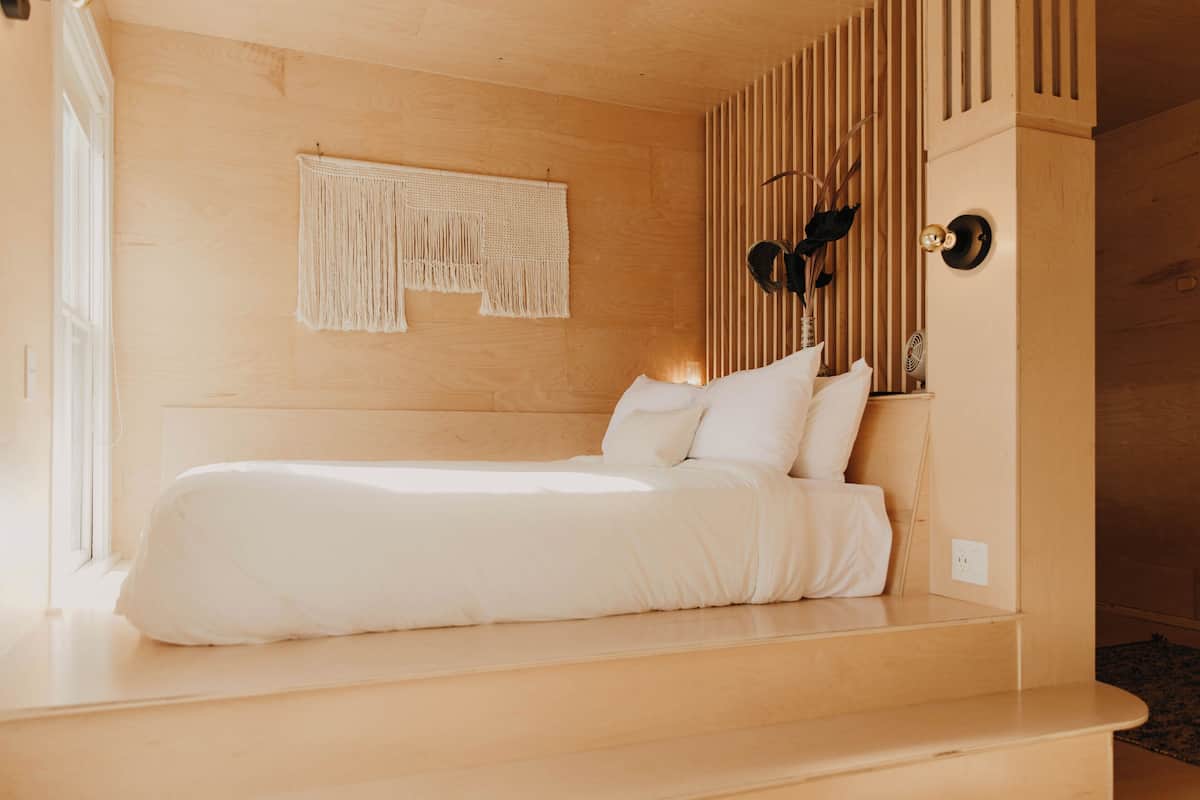 A minimalist bedroom with a wooden headboard, a macrame wall hanging, and a black decorative item on the wall.