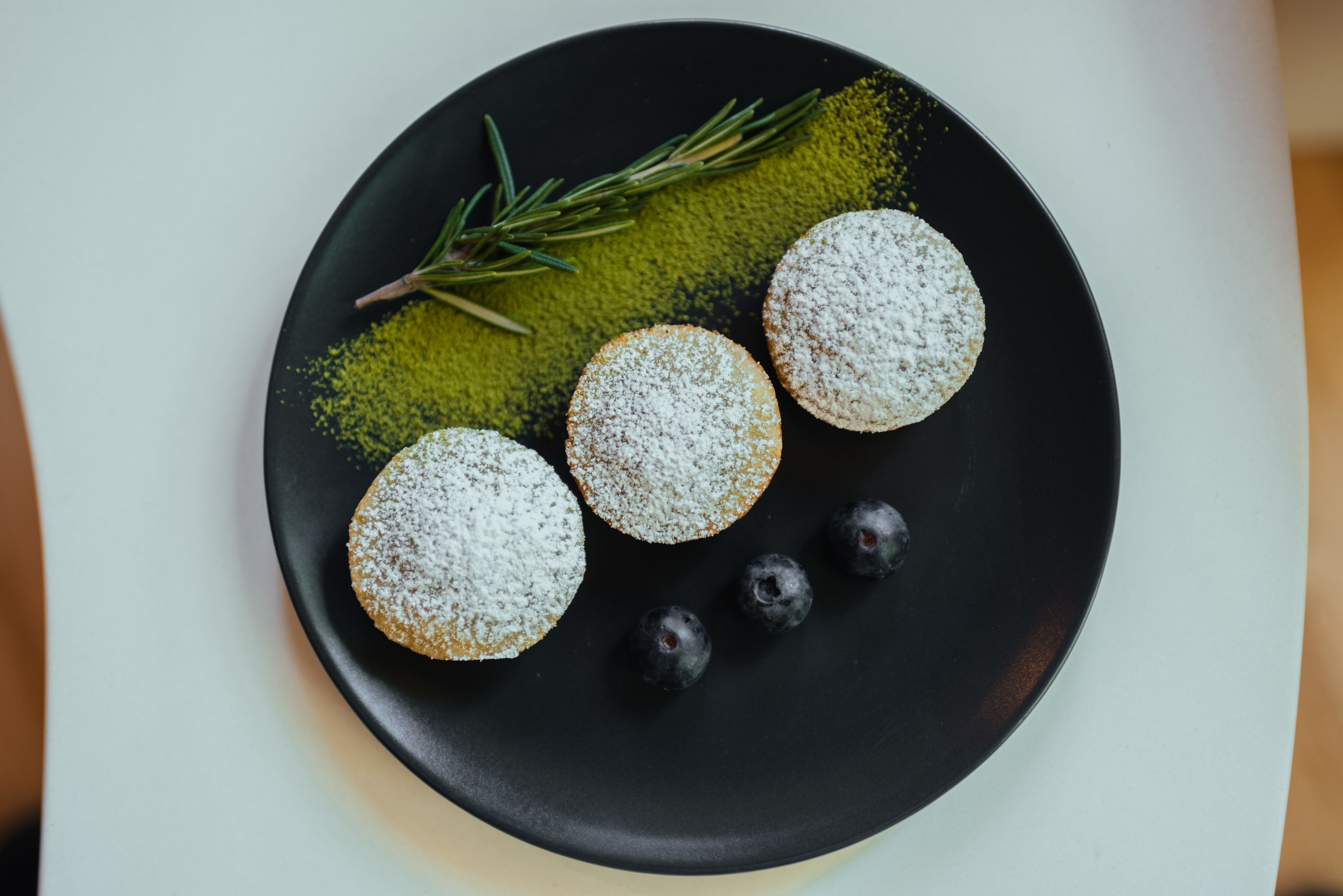 A plate with three round pastries dusted with powdered sugar, accompanied by blueberries and a sprig of rosemary.