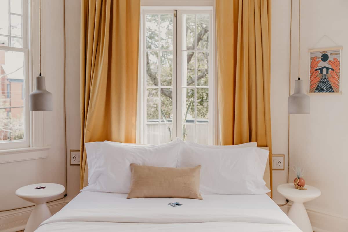 A neatly made bed with white linens and a beige pillow, flanked by two bedside tables with lamps, in a room with a large window and curtains.