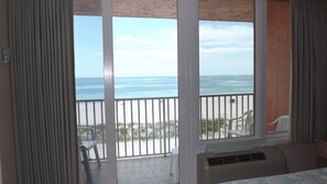 Habitación con vista a la playa y al océano a través de amplios ventanales. Hay dos sillas blancas afuera de la puerta.