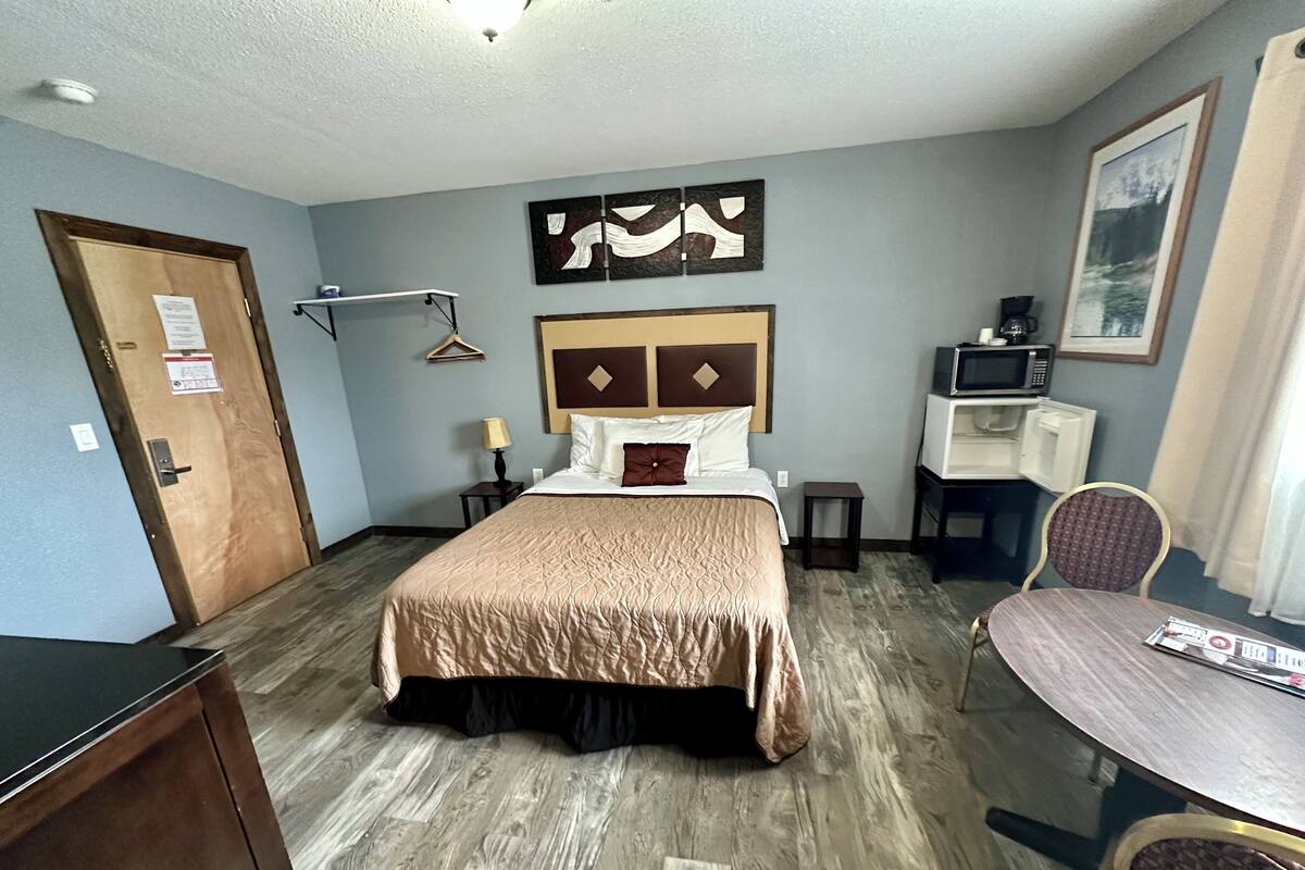 A hotel room with a bed, a desk, a chair, a table, a microwave, and a painting on the wall.