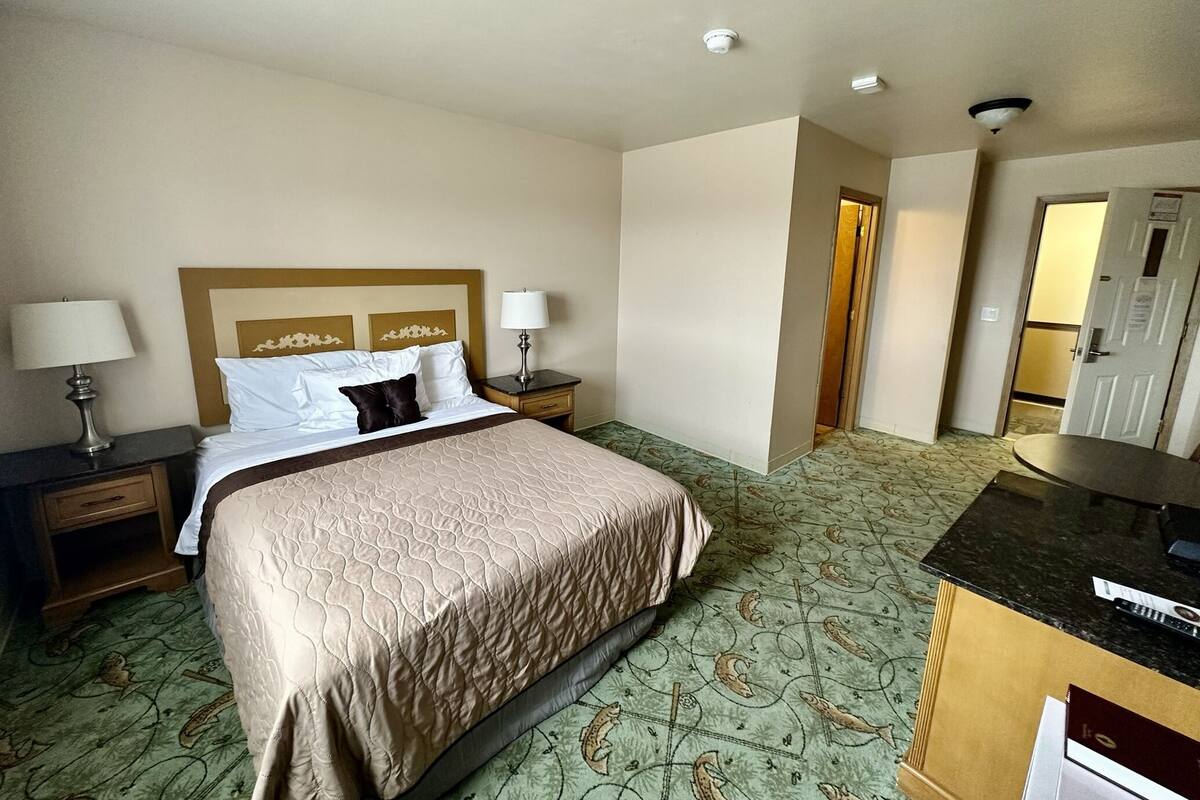 A hotel room with a bed, two bedside tables with lamps, a small desk with a chair, and a patterned carpet.