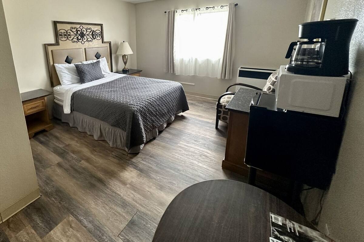 A hotel room with a bed, bedside table, lamp, window with curtains, and a coffee maker.