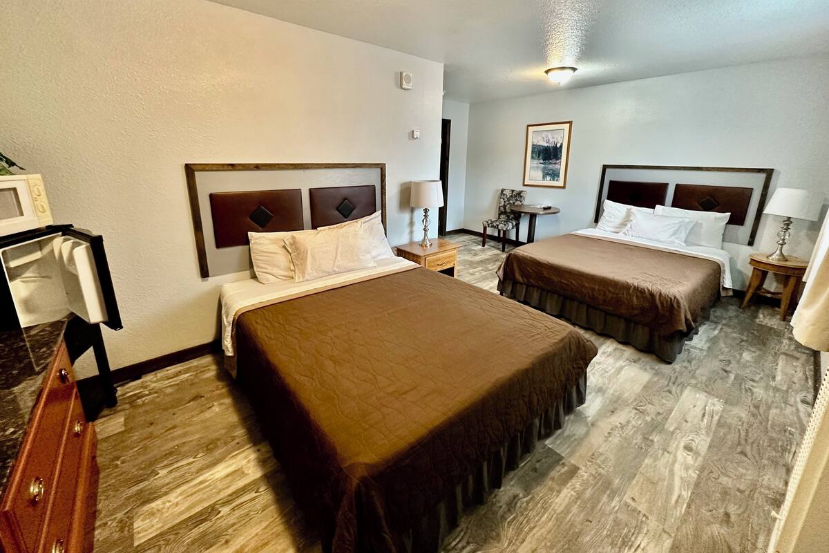 A hotel room with two beds, a desk, a chair, and a small table with a lamp.