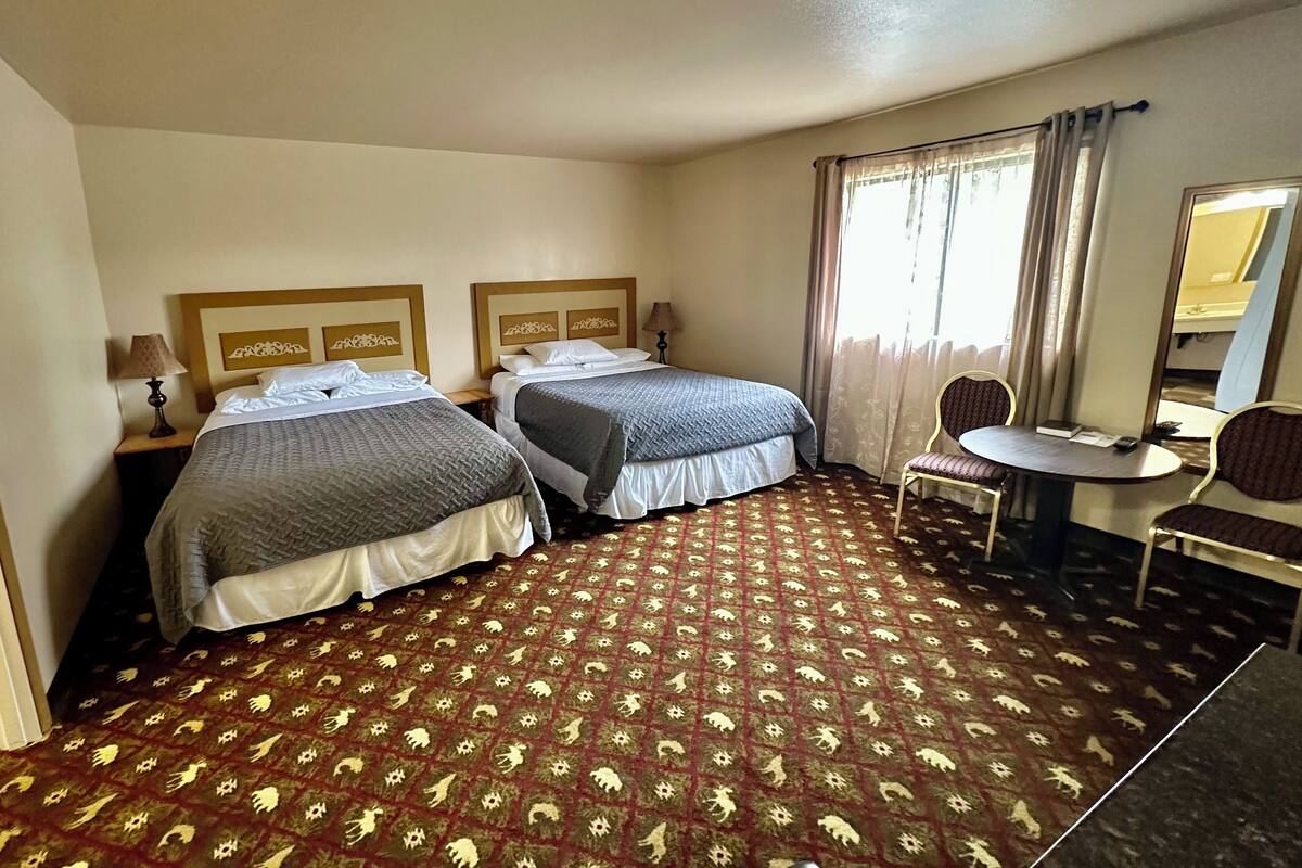 A hotel room with two beds, a small round table with a chair, a mirror, and a patterned carpet.