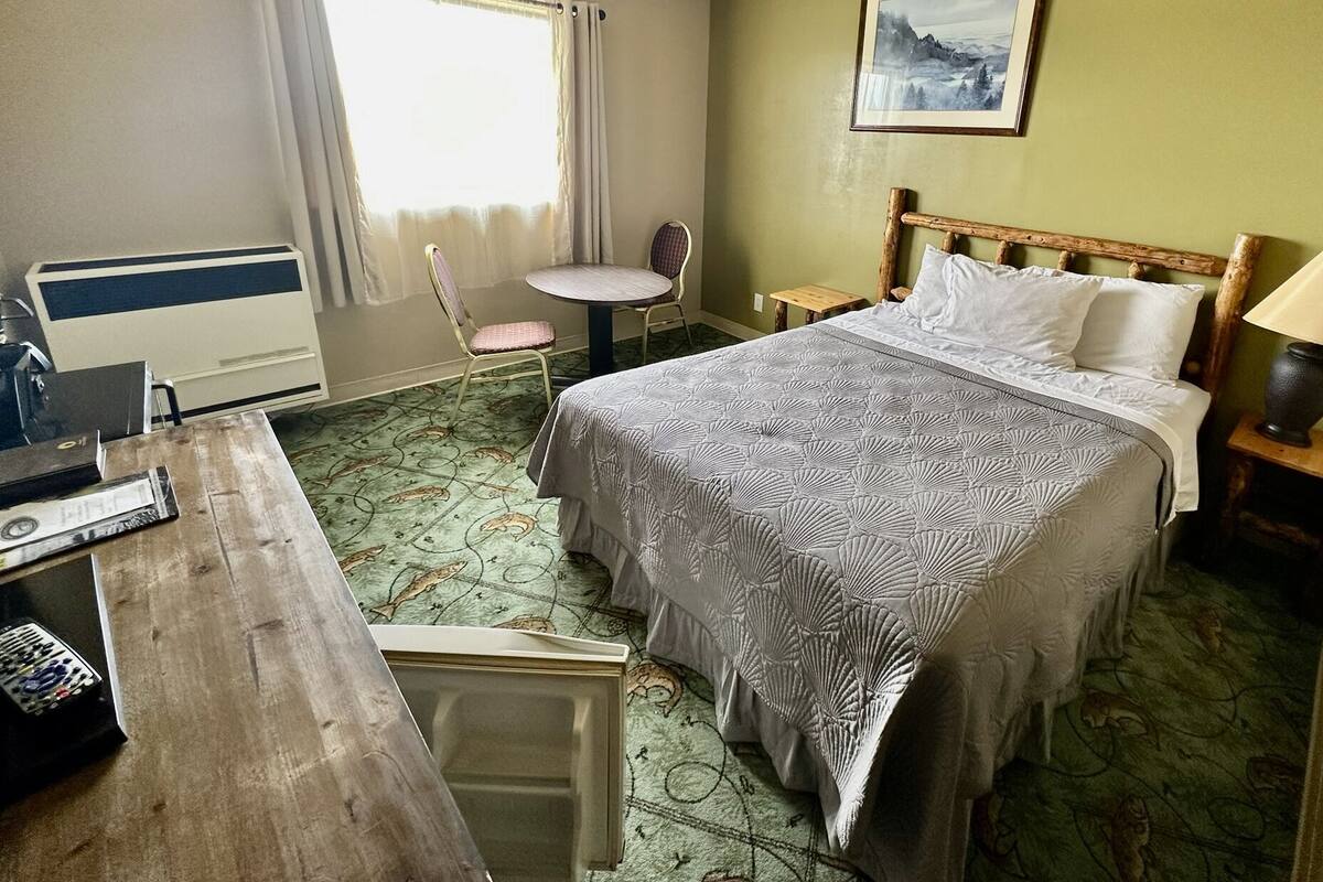 A hotel room with a bed, a desk, a chair, a small table, and a lamp.