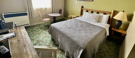 A hotel room with a bed, a desk, a chair, a small table, and a lamp.