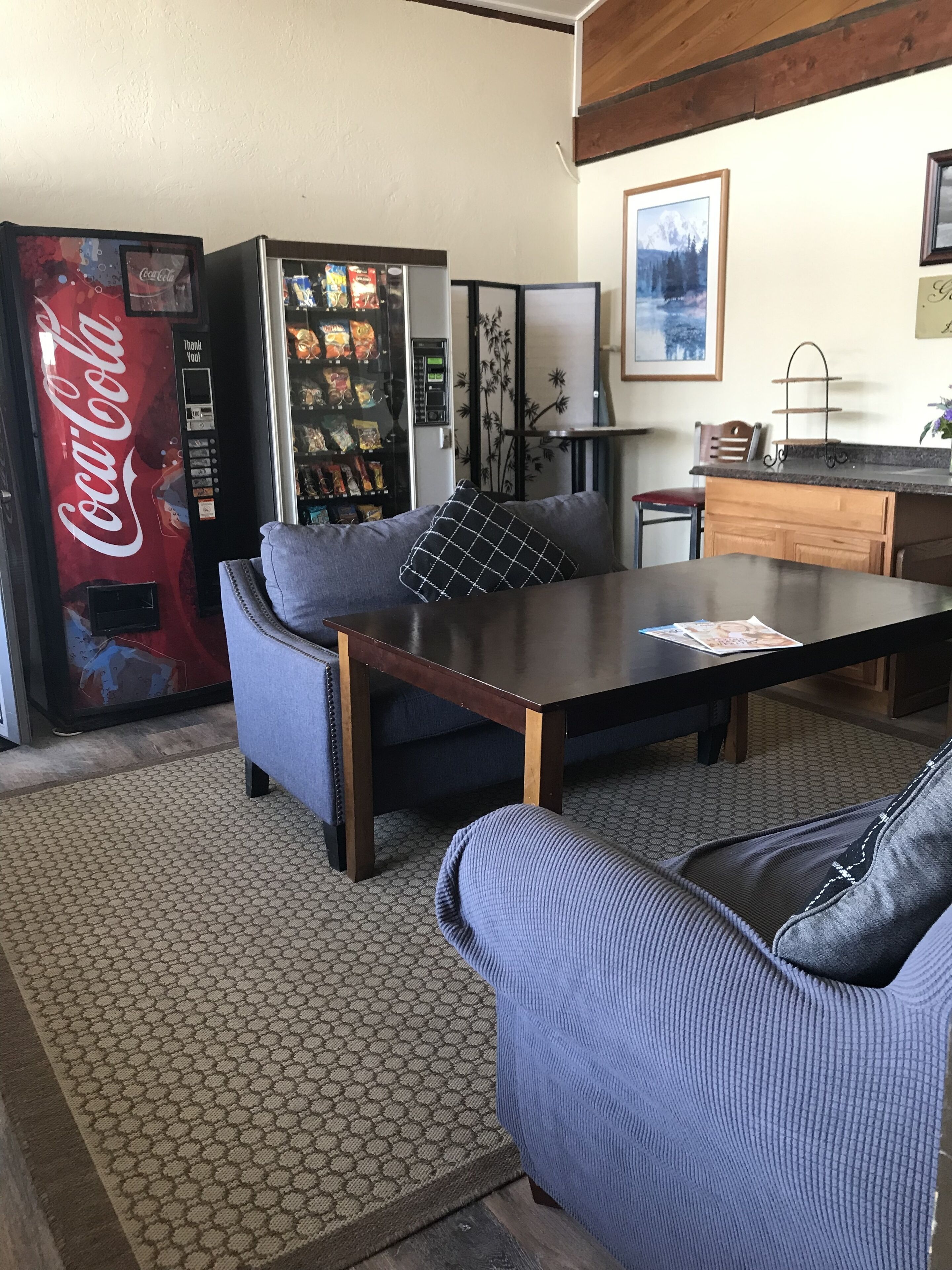 A living room with a sofa, a coffee table, and a kitchen area with a refrigerator and a Coca-Cola vending machine.