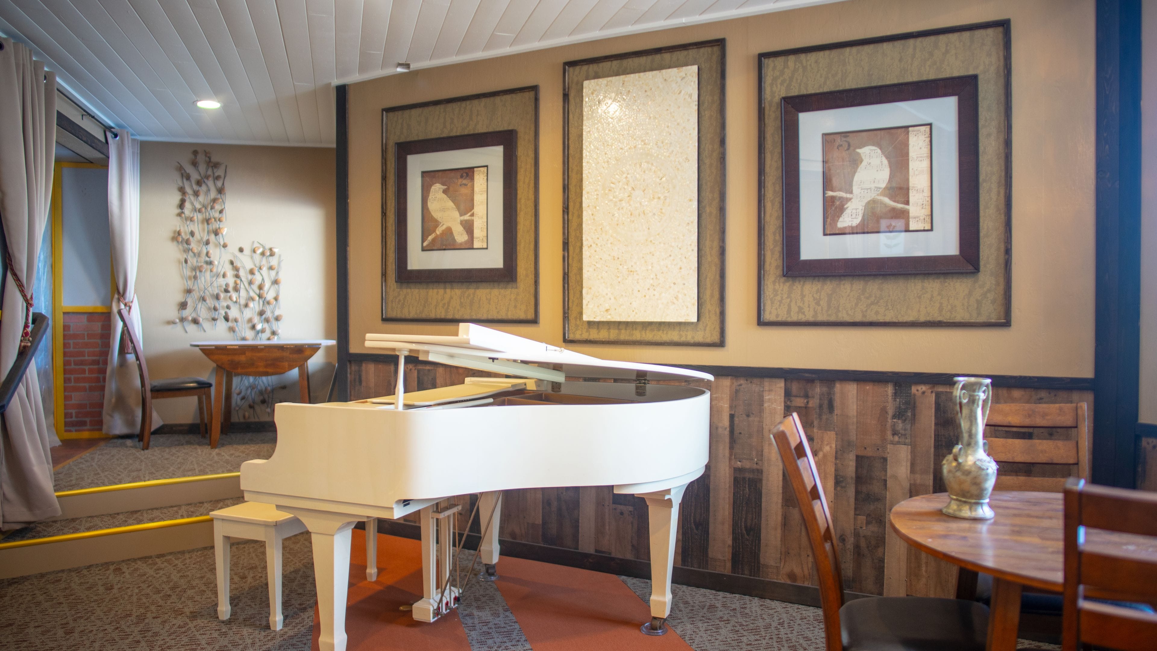 A room with a grand piano, framed artwork on the walls, a wooden bench, and a table with a vase.