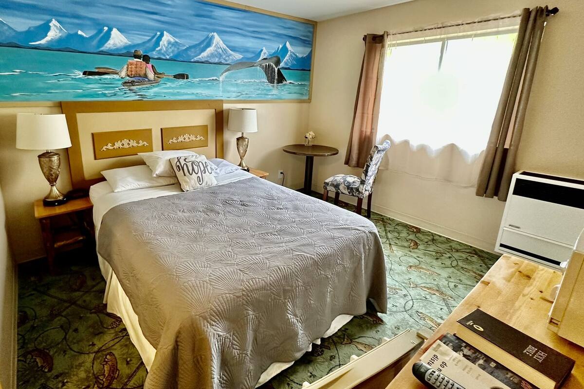 A hotel room with a bed, a painting of a boat on water, a chair, a table, and a desk with a computer.