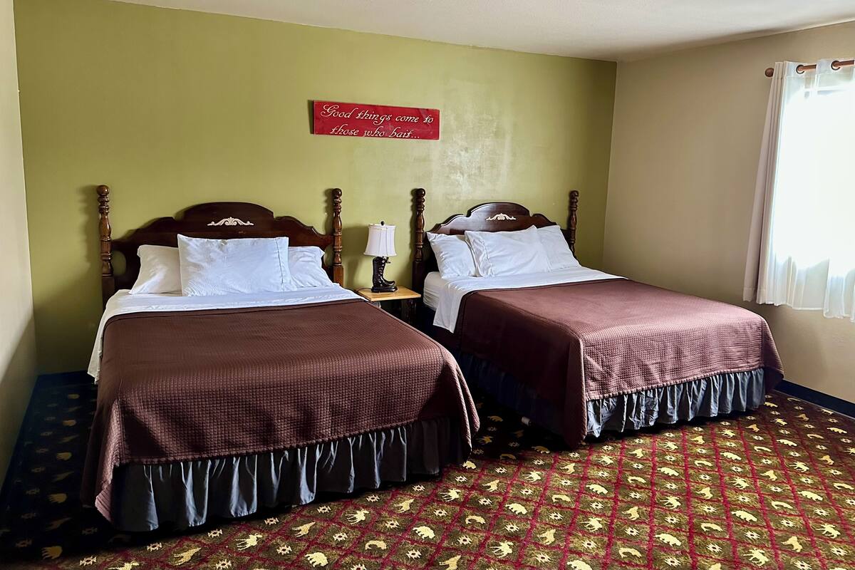 A hotel room with two beds, each with a wooden headboard, white pillows, and a brown bedspread. A red sign on the wall reads "Good things come to those who wait." The room has a patterned carpet and a window with white curtains.