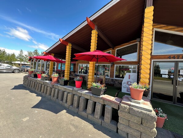A building with a wooden exterior, a covered outdoor seating area with red umbrellas, and a stone wall with potted plants.