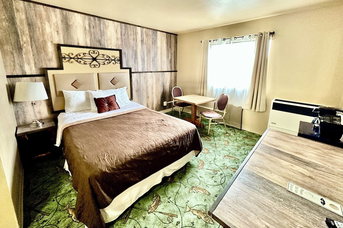 A hotel room with a large bed, a desk, two chairs, a lamp, and a radiator.